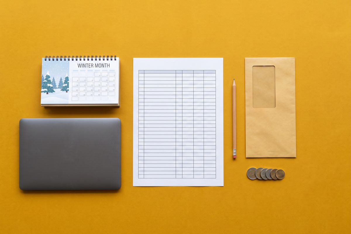 Overhead view of a blank ledger, laptop, envelope, coins, and calendar on a mustard background.
