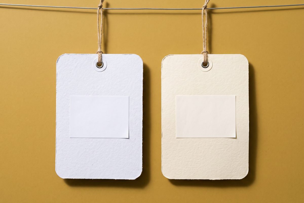 Two oversized blank paper price tags hanging side by side on a plain mustard background