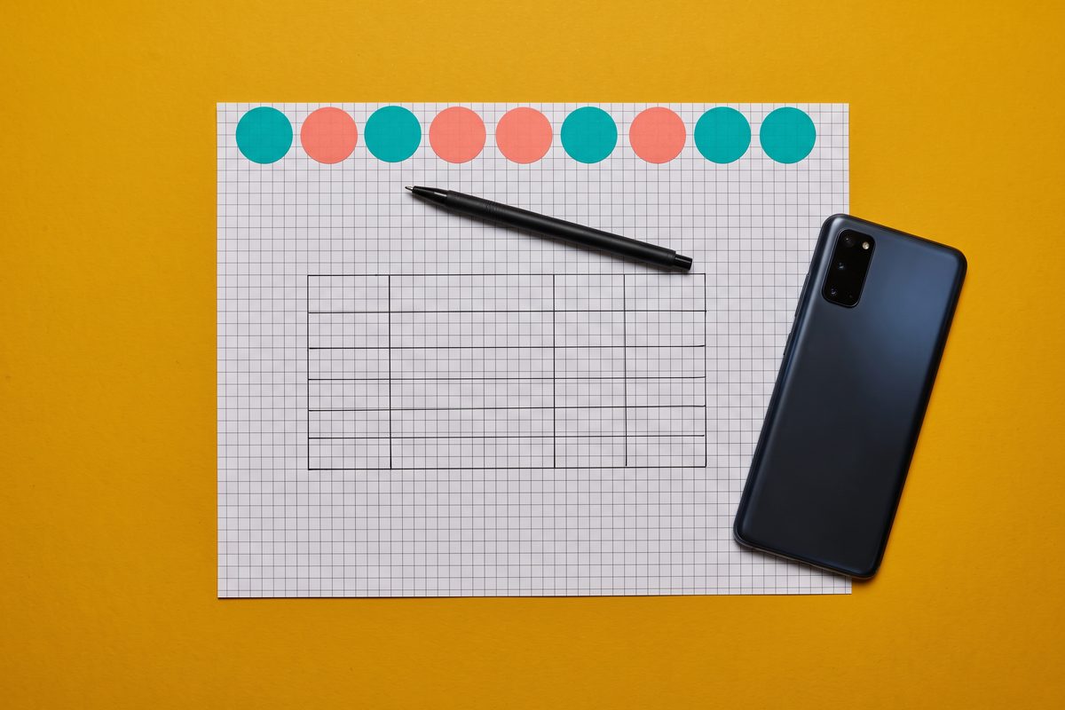 Grid paper with an empty hand-drawn table, a pen, a face-down phone and nine colored stickers on a mustard background