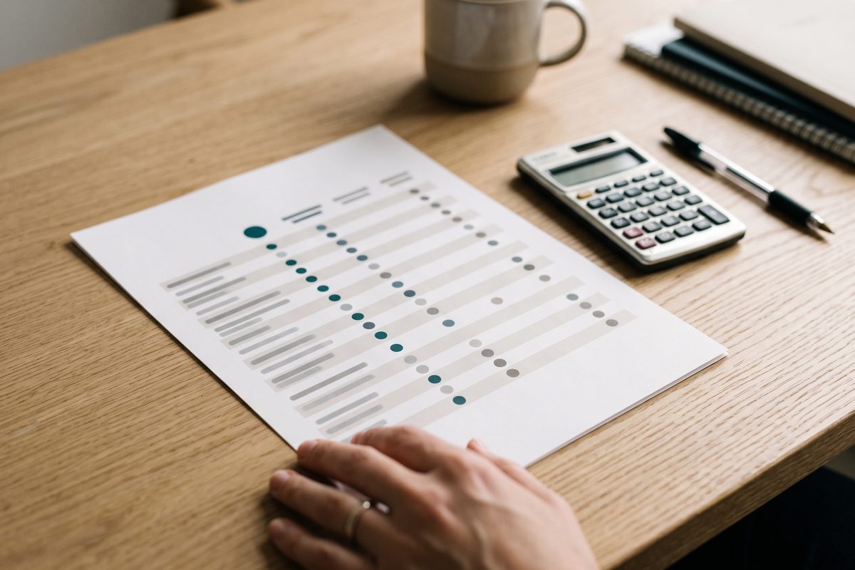 A printed pricing comparison sheet on a wooden desk beside a calculator and pen