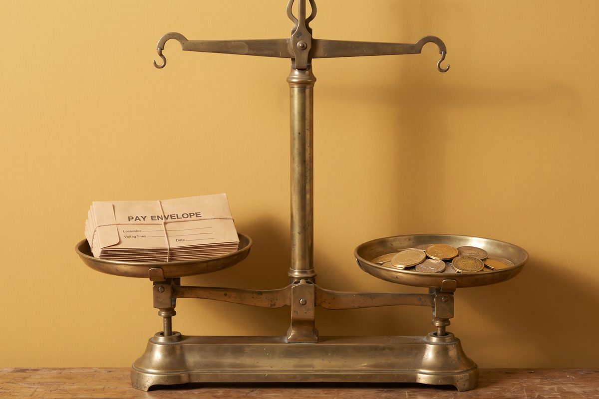 Oversized brass balance scale weighing a stack of pay envelopes against a pile of gold coins on a mustard background.